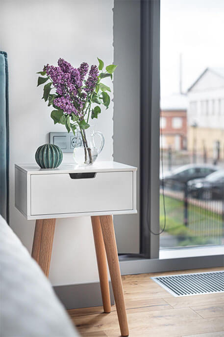 White Bedside Table With Glass Vase Lilac Against White Wall